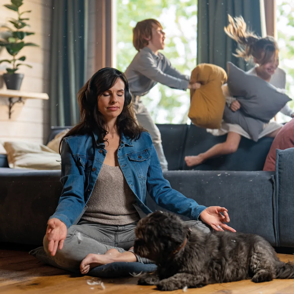Campagnebeeld Aevitae: ontspannen model met koptelefoon in kleermakerszit, op de achtergrond kinderen springend op de bank